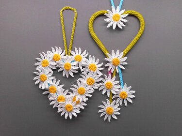 Two crocheted heart decorations with white and yellow daisies on a grey background