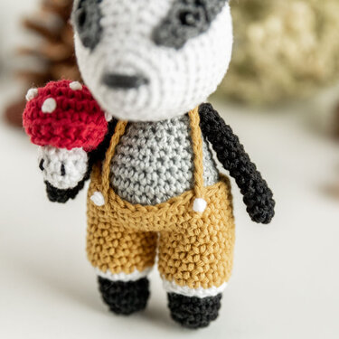 Gehäkeltes Amigurumi‑Tier in senffarbener Latzhose hält roten Pilz