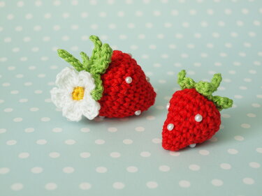 Zwei Mini-Erdbeeren aus rotem Garn mit grünen Blättern und Perlen auf hellblauem, gepunktetem Hintergrund