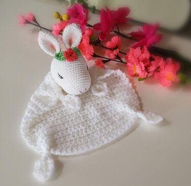 Weißes gehäkeltes Schmusetuch mit Hasenkopf, geschlossenen Augen und roter Blüte, dekoriert mit rosa Blumenzweigen.