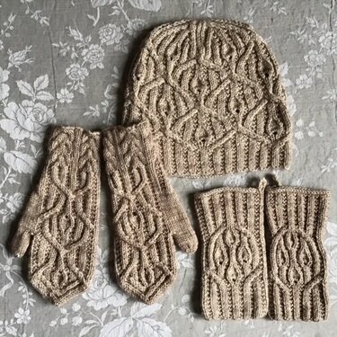Beiges Strickset mit Zopfmuster: Mütze, Fäustlinge und Pulswärmer auf gemusterter Oberfläche