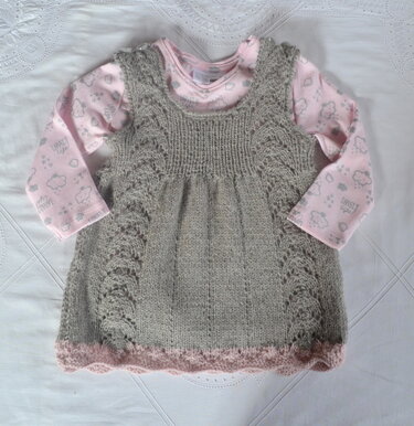 Grau gestricktes Babykleid mit Lochmuster an den Seiten und rosa gewelltem Saum, darüber ein rosa Langarmshirt mit Wolkenmotiven
