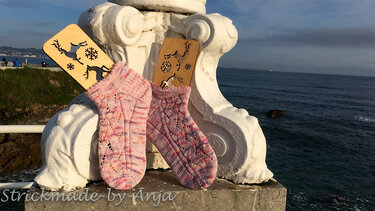 Zwei gestrickte Damensocken in Rosa- und Violetttönen mit Zopf- und Lochmuster auf weißem Steinsims vor dem Meer, mit dekorativen Holzschildern im Hintergrund