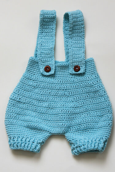 Hellblauer gehäkelter Baby-Strampler mit Trägern und dunklen Knöpfen, liegend.