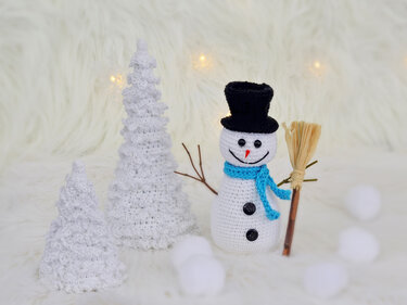 Gehäkelter Schneemann mit schwarzem Zylinder, blauem Schal und zwei weißen Häkelbäumen auf hellem Untergrund