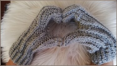 Zwei Hände in grauen gehäkelten Stulpen formen ein Herz auf weißem, flauschigem Untergrund.