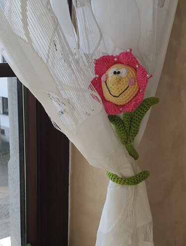 Gehäkelter Blumen-Raffhalter in Pink mit gelbem Smiley-Gesicht und grünen Blättern an weißem Vorhang