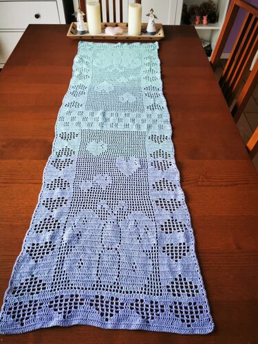 Langer gehäkelter Tischläufer in Türkis‑Lavendel mit Filetmuster auf einem Holztisch, mit Kerzenhalter am oberen Ende.