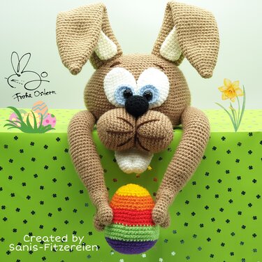 Brauner gehäkelter Osterhase mit schlaffen Ohren und großen Augen hält ein bunt gestreiftes Regenbogen-Osterei.