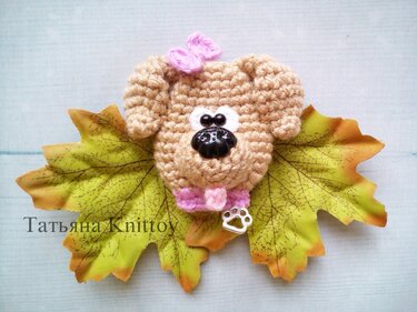 Nahaufnahme eines gehäkelten Hundekopfes in Beige mit rosa Schleife und Pfotenanhänger auf Herbstblättern