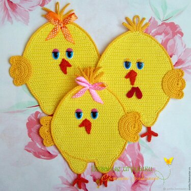 Drei gelbe gehäkelte Küken mit applizierten Augen, orange-roten Schnäbeln, kleinen Schleifen und Flügeln auf floralem Hintergrund.