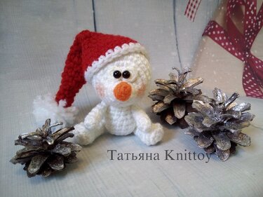 Weißer gehäkelter Schneemann mit roter Weihnachtsmütze, oranger Nase und schwarzen Perlenaugen neben silberbemalten Tannenzapfen und Geschenkverpackung