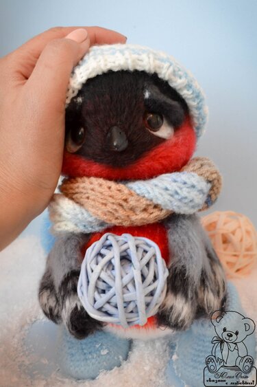 Gehäkelter Amigurumi-Vogel mit weiß-blauer Mütze und Schal, hält eine Garnkugel; eine Hand streichelt den Kopf