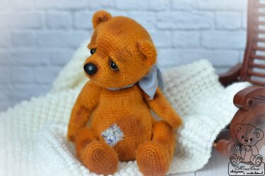 Gehäkelter orangebrauner Teddybär mit hellblauem Halstuch und floralem Patch auf einer weißen Decke vor einer Ziegelwand.