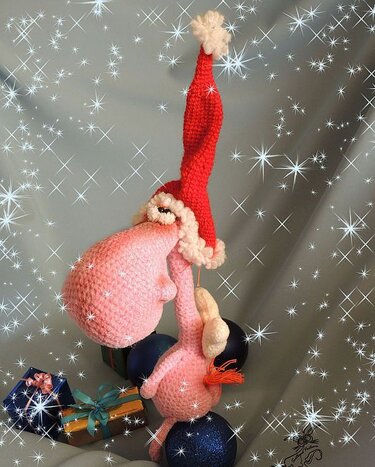 Rosa gehäkelter Amigurumi-Flamingo hängt kopfüber an roter Weihnachtsmütze vor funkelndem Hintergrund mit Kugeln und Geschenken.