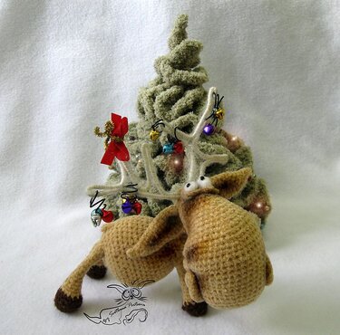 Braunes Amigurumi-Rentier mit geschmücktem Geweih vor kleinem grünen Häkelbaum.