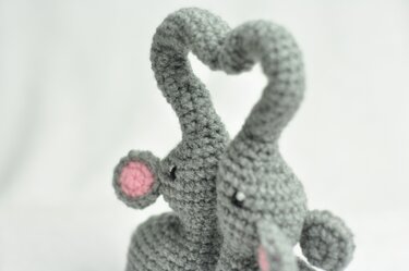 Grey amigurumi elephants with trunks forming a heart, showing stitch texture and pink ear detail