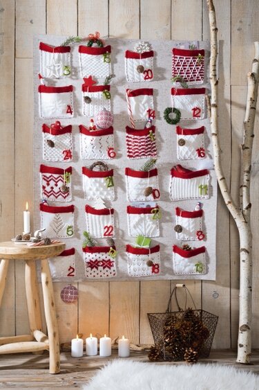 Gestrickter Adventskalender mit 24 roten und weißen Taschen an einer Holzwand, daneben Kerzen und Tannenzapfen.