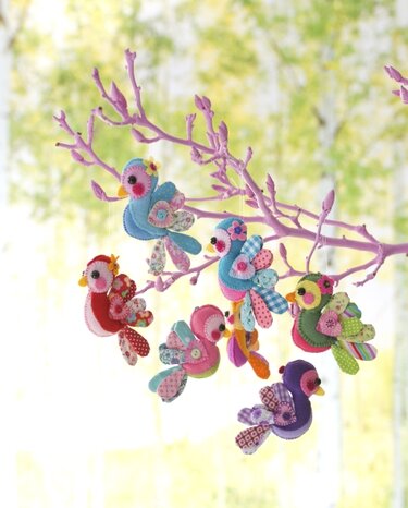 Mehrere bunte, genähte Filzvögel mit Stoffmustern hängen an einem rosa Ast vor hellgrünem Hintergrund.