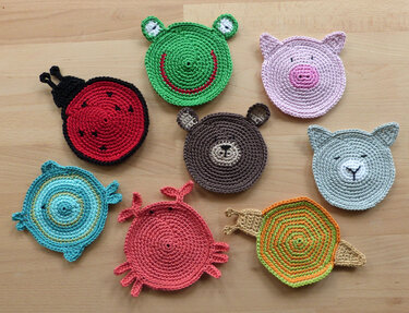 Acht bunte, gehäkelte Untersetzer in Tierformen auf einem Holztisch: Frosch, Schwein, Marienkäfer, Bär, Katze, Schildkröte, Krabbe und Schnecke.