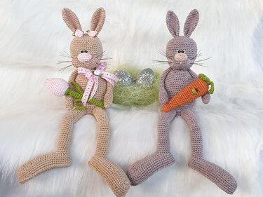 Zwei gehäkelte Amigurumi-Hasen mit Karotte und Blume vor Osternest auf weißem Fell