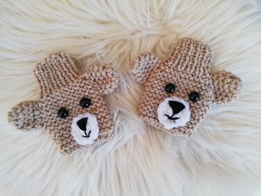 Zwei beige gestrickte Baby-Fäustlinge mit Bärengesichtern auf weißem Fell