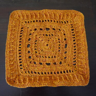 Gehäkeltes Granny-Quadrat in Senfgelb mit schimmerndem Garn und Lochmuster auf dunklem Grund.