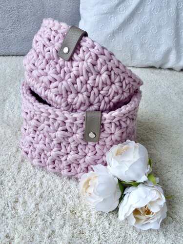 Zwei pastellrosa gehäkelte Körbe ineinander gestellt mit braunen Kunstledergriffen auf hellem Teppich neben weißen Blumen.