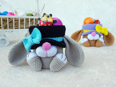 Zwei gehäkelte Hasenkörbchen mit Hüten und Schleifen, eines grau im Vordergrund und eines braun im Hintergrund, beide gefüllt mit österlicher Dekoration.