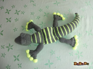 Gehäkelte Eidechse in Grau mit neongelben Streifen und Zehen auf hellgrünem Stoff mit Blattmuster.