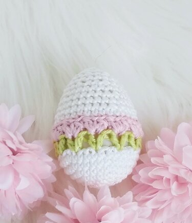 Nahaufnahme eines gehäkelten Oster-Eis in Weiß mit rosa und hellgrünen Streifen, umgeben von rosa Stoffblüten auf weißem, flauschigem Hintergrund.