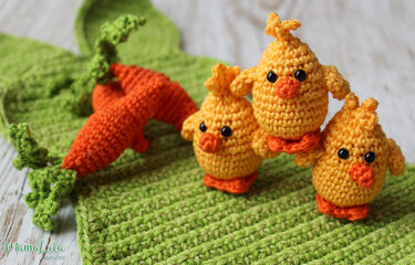 Drei gelbe gehäkelte Amigurumi-Küken mit orangefarbenen Schnäbeln neben zwei gehäkelten Karotten auf grünem Häkelstoff