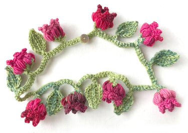 Gehäkelte Blumenranke mit rosa und magentafarbenen Blüten, grünen Blättern und kleinem Knopf auf weißem Hintergrund.