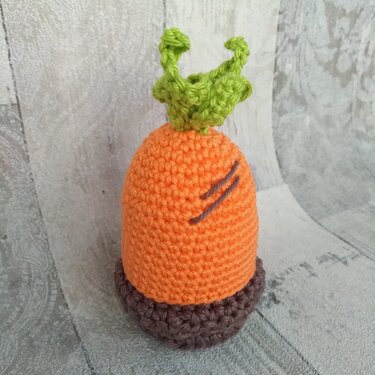 Gehäkelter Karotten-Eierwärmer mit grünen Blättern und braunem Sockel auf hellem Hintergrund.