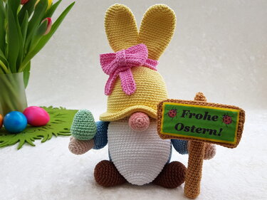 Gehäkelter Amigurumi-Wichtel mit gelbem Hasenhut, rosa Schleife, Schild 'Frohe Ostern' und kleinem Ei