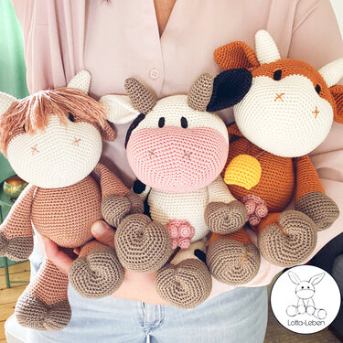 Three crocheted amigurumi cow plush toys held by a person: shaggy brown, white-and-black spotted with a pink muzzle, and orange-brown.