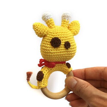 Yellow crocheted giraffe teether with brown spots, white-tipped ossicones, red scarf and wooden ring held by a hand on a white background.