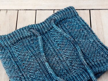 Gestrickter Loopschal in Blau-Grau mit Fischgrätmuster und Kordelzug auf hellem Holz