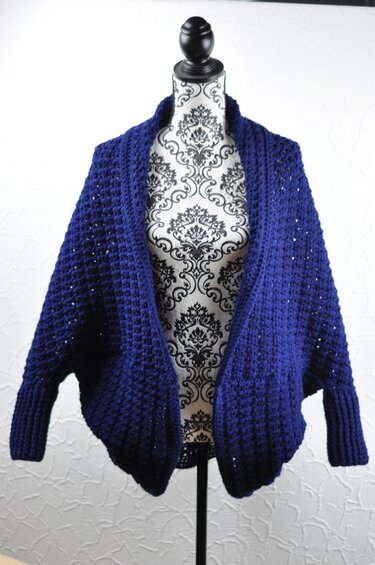 Dark blue crocheted open-front shrug with textured stitch and ribbed cuffs on a mannequin.