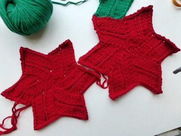 Zwei rote, handgestrickte Sterne mit sichtbarem Muster auf weißer Fläche, daneben ein grünes Wollknäuel.