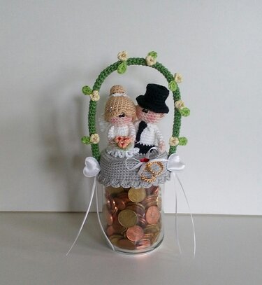 Gehäkeltes Brautpaar mit Zylinder und Schleier auf einem mit Münzen gefüllten Glas, dekoriert mit einem grünen Blumenbogen.