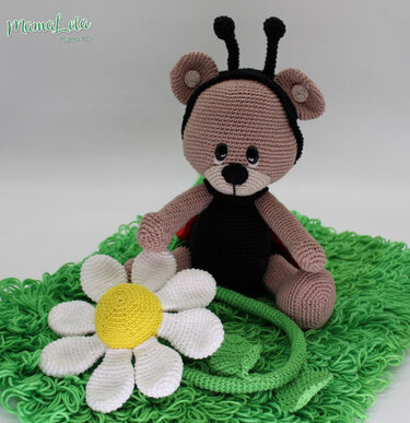 Amigurumi-Teddybär mit Marienkäfer-Antennen neben gehäkelter Margerite auf grünem Garn