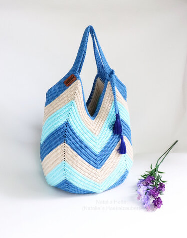 Gehäkelte Henkeltasche mit Chevron-Zickzack in Blau-, Türkis- und Beigetönen, zwei Quasten und langen Henkeln, neben lila Blumen