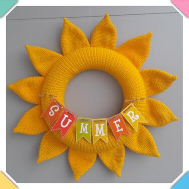 Gehäkelter gelber Sonnenblumenkranz mit Wimpelkette SUMMER in der Mitte, an einer grauen Wand hängend.