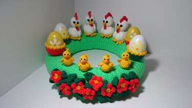 Gehäkelter grüner Osterkranz mit vier weißen Hühnern, vier gelben Küken, bunten Eiern in Körbchen und roten Blüten.