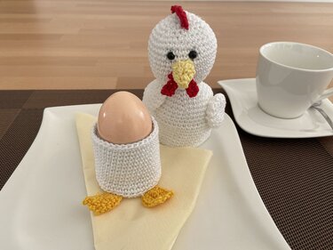 Weiß gehäkeltes Huhn neben einem Ei im gehäkelten Eierbecher mit gelben Füßen auf einem Teller