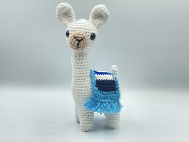 White crocheted llama amigurumi with navy and turquoise saddle and fringe, standing on plain background