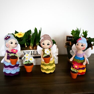 Drei gehäkelte Feenpuppen mit Flügeln, jede hält einen kleinen Blumentopf mit gehäkelter Blume, vor Topfpflanzen