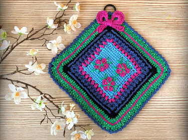 Farbenfroher gehäkelter Topflappen mit Blumenmotiven, auf Holzuntergrund neben Blütenzweig liegend