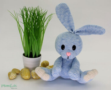 Hellblauer gehäkelter Amigurumi-Hase mit langen Ohren und rosa Herznase, sitzend neben Topfgras und goldfarbenen Deko-Steinen.
